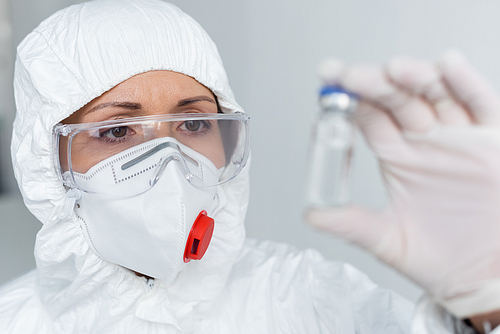 Scientist in protective mask and goggles holding vaccine on blurred foreground