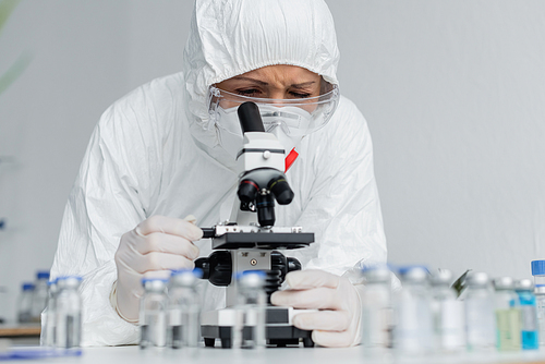 Scientist in goggles and hazmat suit looking at microscope near vaccines on blurred foreground