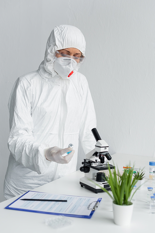 Scientist in hazmat suit looking at vaccine near clipboard and microscope in laboratory