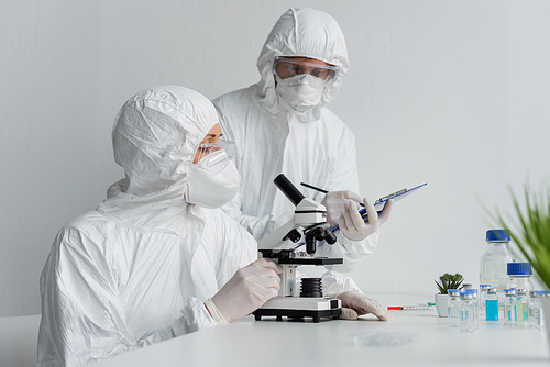 Scientist using microscope near colleague with clipboard and vaccines