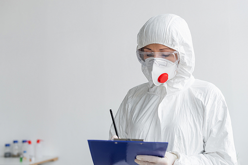 Scientist in medical mask and protective suit writing on clipboard on blurred foreground
