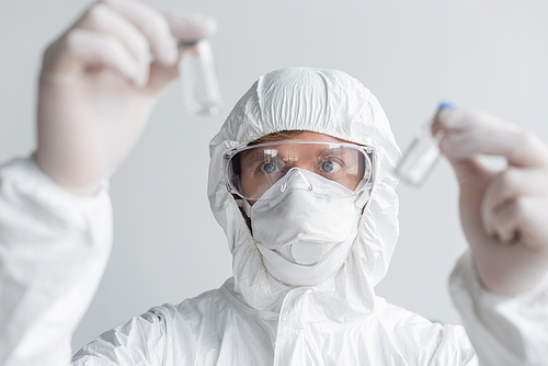Scientist in hazmat suit looking at vaccines on blurred foreground