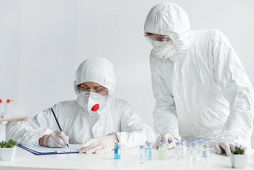 Scientist writing on clipboard near colleague and vaccines on blurred foreground