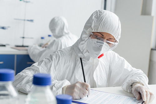 Scientist writing on clipboard near colleague on blurred background in laboratory