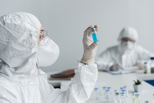 Scientist looking at jar with vaccine near colleague on blurred background