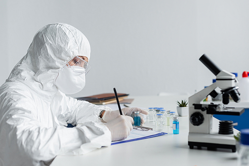 Scientist in protective uniform writing on clipboard near microscope and vaccines