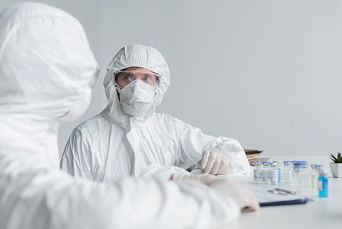 Scientist in hazmat suit looking at colleague near clipboard and vaccines