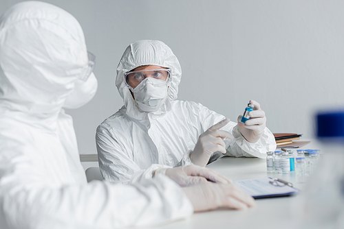 Scientist in protective suit pointing at vaccine near colleague on blurred foreground in laboratory