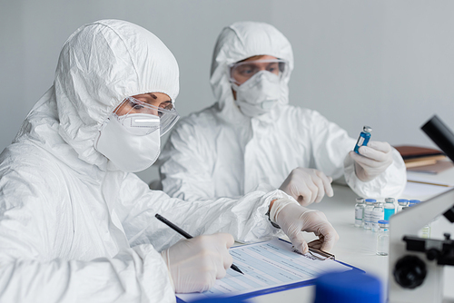 Scientist writing on clipboard near colleague with vaccines on blurred background