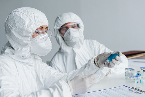 Scientist picking up vaccine in syringe near clipboard and colleague on blurred background