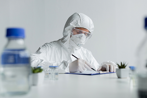 Scientist writing on clipboard near vaccines and plants on blurred foreground