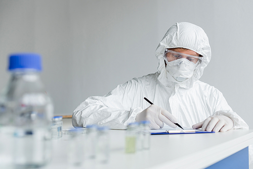 Scientist writing on clipboard near vaccines on blurred foreground on table