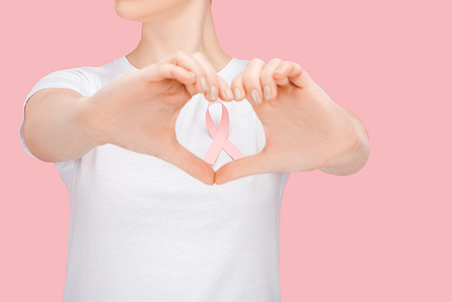 partial view of woman in white t-shirt doing heart sign with hands around pink breast cancer ribbon isolated on pink