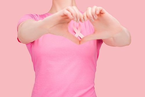 cropped view of woman in pink t-shirt showing heart sign with hands around breast cancer ribbon isolated on pink