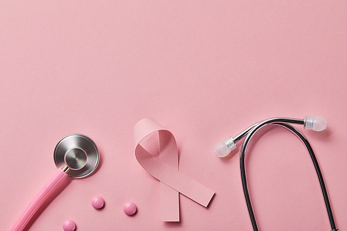 top view of pink metal stethoscope, breast cancer ribbon and three pills on light pink background