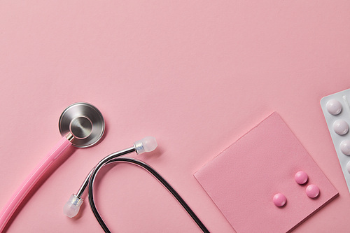 top view of pink stethoscope, three pills on piece of paper and blister pack on light pink background