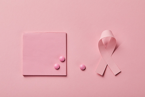 scattered pills on piece of paper near breast cancer ribbon on light pink background