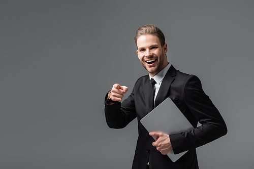 laughing businessman pointing with finger while holding laptop isolated on black