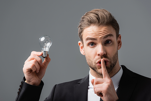 thrilled businessman holding light bulb while showing secret gesture isolated on grey