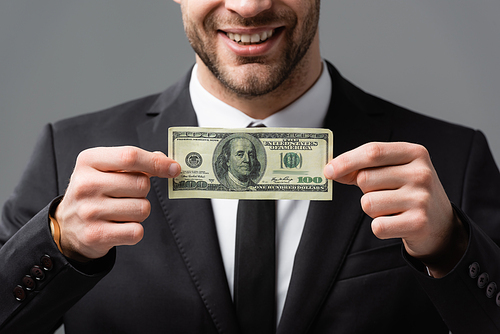 cropped view of smiling businessman holding dollar banknote isolated on grey, blurred background