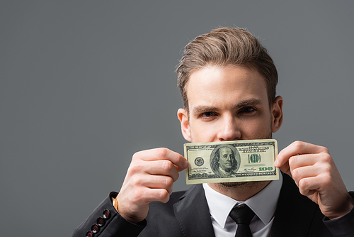 young businessman covering mouth with dollar banknote isolated on grey