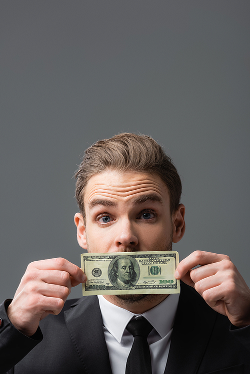 excited businessman covering mouth with dollar banknote isolated on grey