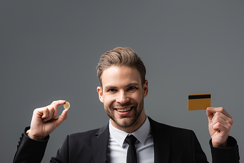 cheerful businessman holding coin and credit card isolated on grey