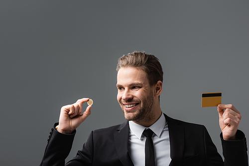 joyful businessman holding credit card and coin isolated on grey