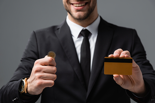 cropped view of smiling businessman holding coin and credit card on blurred background isolated on grey