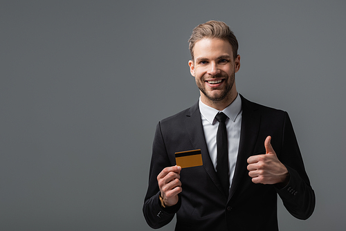 pleased manager showing thumb up while holding credit card isolated on grey
