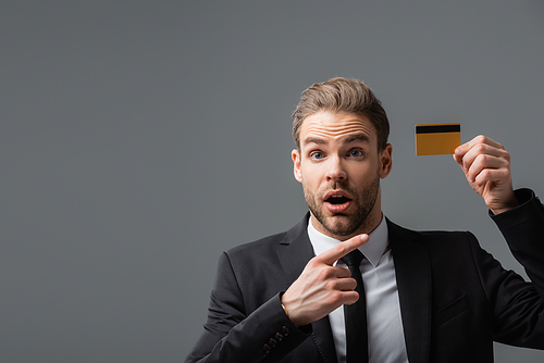 amazed businessman with open mouth pointing at credit card isolated on grey