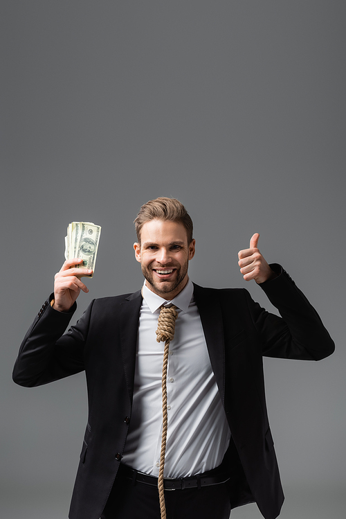 joyful businessman with rope on neck holding money and showing thumb up isolated on grey