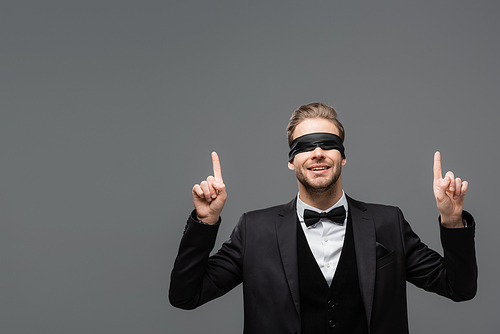smiling businessman in blindfold pointing up with fingers isolated on grey