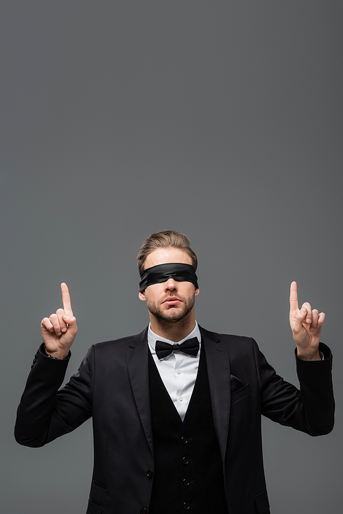 young, blindfolded businessman pointing up with fingers isolated on grey