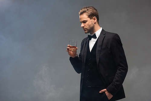 young businessman holding whiskey while standing with hand in pocket on grey background with smoke