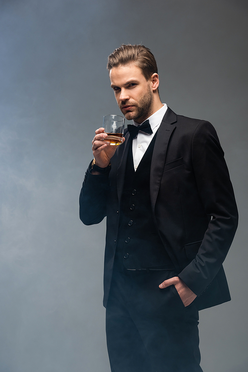 elegant businessman standing with hand in pocket while holding whiskey on grey background with smoke