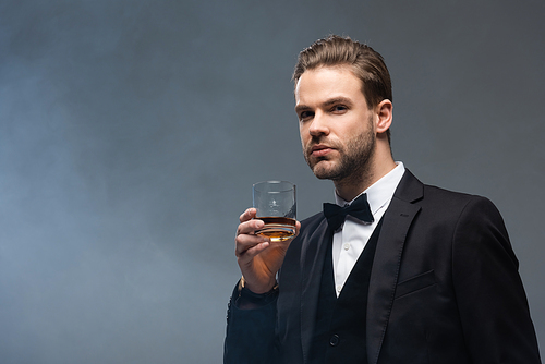 confident businessman holding glass of whiskey on grey background with smoke