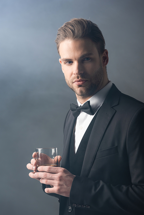 confident businessman looking away while holding glass of whiskey on grey background with smoke