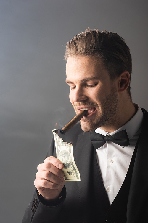 wealthy businessman lighting cigar with hundred dollar banknote on grey background with smoke