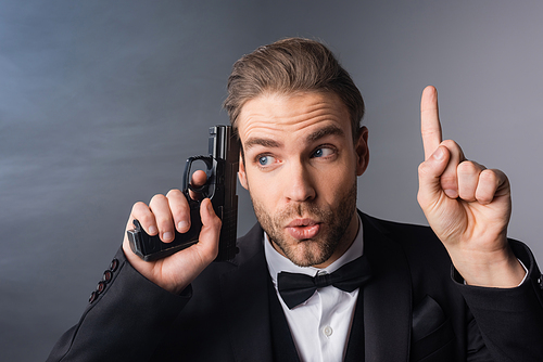 excited businessman showing idea gesture while holding weapon near head on grey background with smoke