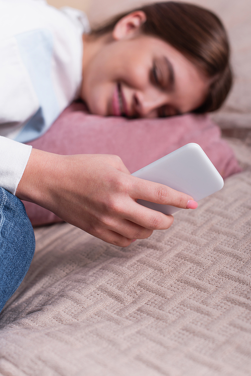 blurred and sad teenage girl holding smartphone while lying on bed