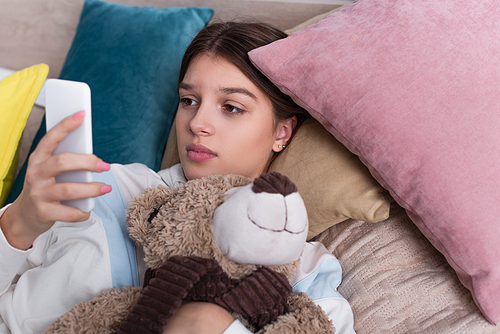 sad teenage girl using smartphone and holding teddy bear