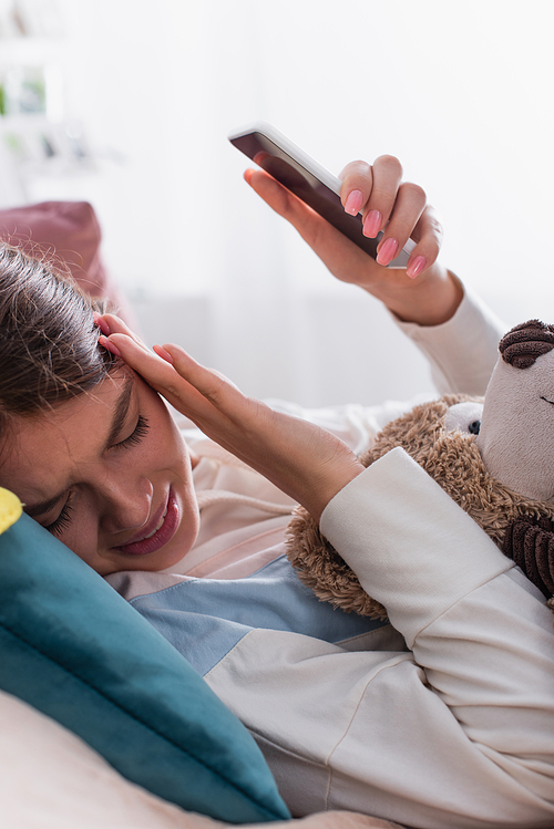 frustrated teenage girl gesturing while using smartphone and lying with teddy bear