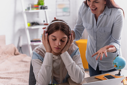 teenage girl in wireless headphones listening music near angry mother screaming on blurred background