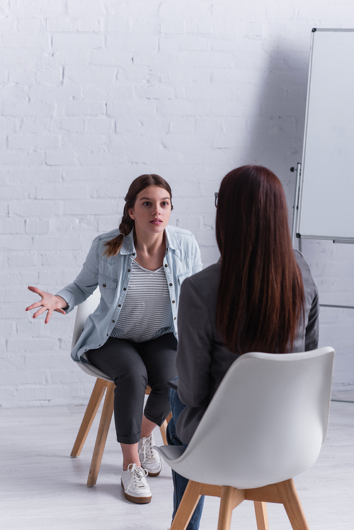 upset teenage girl sitting and gesturing while looking at psychologist on blurred foreground