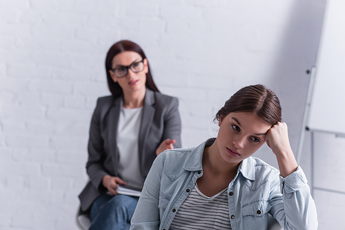 worried teenage girl looking away with psychologist sitting behind on blurred background