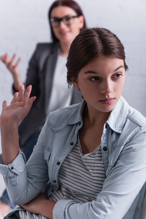 depressed teenage girl looking away and gesturing near psychologist sitting behind on blurred background