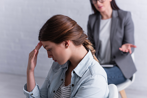 depressed teenage girl looking away near psychologist sitting behind on blurred background
