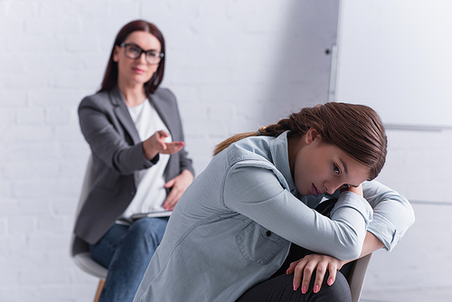 frustrated teenage girl looking away with psychologist sitting behind on blurred background