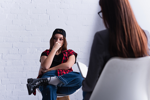 sad teenage patient in cap sitting and covering mouth near psychologist on blurred foreground